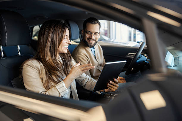 Happy buyer and female car dealer sitting in classy car at car salon and smiling at tablet.