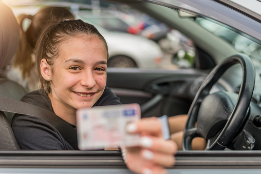 Caucasian Female teen  sitting behind wheel and holding out his driving license through car window - new drivers concept. Travel and rental concept. Copy space, close up